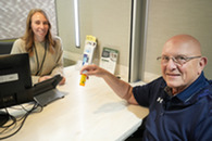 a person with long hair sitting behind a desk and bald person wearing glasses sitting in front of desk holding a pill bottle 