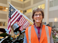 a person with brown short curly hair wearing glasses and a reflective vest standing in front of flags 