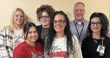 a group of five women and one man standing in two rows  in front of a tan wall all smiling 