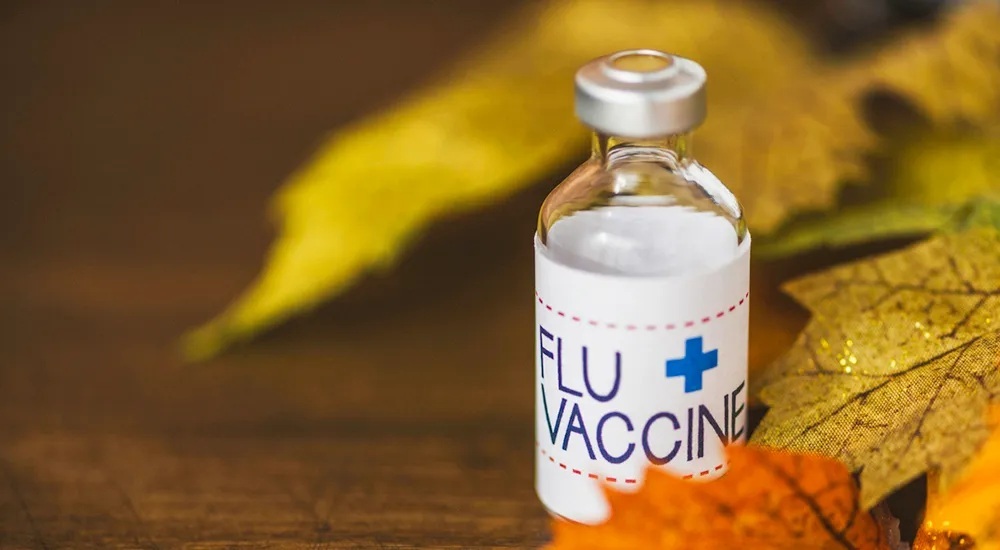 Close-up of a flu vaccine vial surrounded by autumn leaves on a wooden surface.