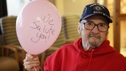 Elderly man wearing a Navy cap and red hoodie holding a pink balloon that says 'We salute you!' 