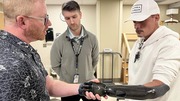 Three men in a clinical setting, one wearing a black carbon fiber prosthetic arm, engaged in conversation.