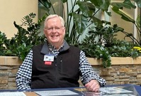 older person with white hair wearing glasses and vest sitting behind a table 