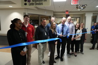 a large group standing behind a blue ribbon and cutting ribbon with scissors 
