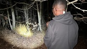 Person wearing a dark hoodie shining a flashlight into a dense forest at night.