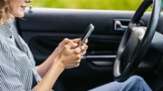 Woman sitting in driver's seat of car, typing on smartphone with both hands near steering wheel.