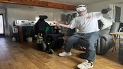 Army Veteran Jeffrey Brunnelson holding two controllers, posing dynamically next to a motorized wheelchair in a living room.