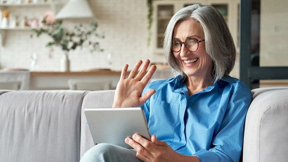 Smiling older woman waving at a tablet while sitting on a couch in a cozy living room.