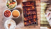 Grilled skewers of marinated meat cooking on a small tabletop grill with bowls of rice, peanut sauce, and spicy chili sauce on the side.