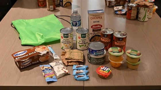 Assortment of canned goods, boxed chickpea rotini, brown rice, snacks, and bottled water on a wooden table.