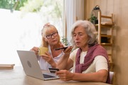 Two elderly women using a laptop for online shopping, one holding a credit card and speaking into a smartphone.