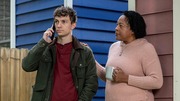 Young man in a green jacket talking on a phone while a woman in a pink sweater gently touches his shoulder outside a house.