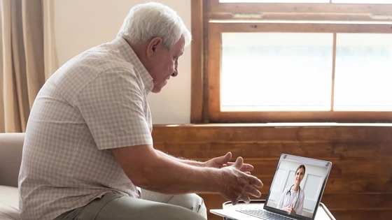 Elderly man sitting by a window engaged in a video call with a female doctor on a laptop screen.