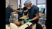 Medical team in scrubs and masks performing a procedure on a patient's head with monitoring equipment nearby.