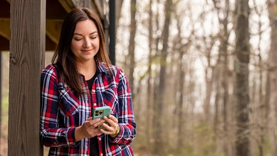 Young woman in a red plaid shirt smiling while using her smartphone outdoors near wooden structure in a leafless forest.