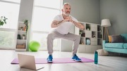 Older man exercising indoors, squatting on a purple mat with a laptop open and a green exercise ball in the background.
