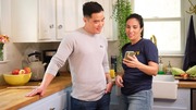 A man and woman standing in a kitchen, looking at a smartphone together and smiling.