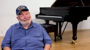 Daniel Bremer sitting and smiling near a black grand piano in a bright room.
