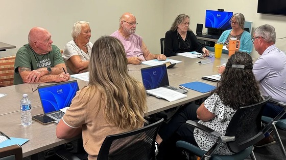 Seven adults engaged in a focused meeting around a conference table with laptops and documents.