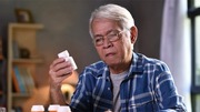 Elderly man in glasses examining a white pill bottle in a cozy, softly lit room.