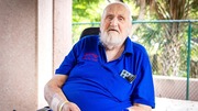 Elderly man wearing a blue polo shirt sitting outdoors near a fence and palm trees.