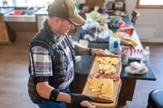 Man wearing a cap and plaid shirt holding a wooden board with an assortment of cheeses, crackers, and sliced meats in a kitchen.