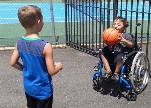 Two young boys, one of them a wheelchair user, play adaptive basketball.