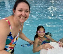 A girl hanging from the wall of a pool posing with her mother.
