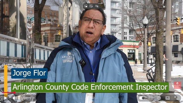 A man in a blue jacket faces the camera and speaks. He is surrounded by streets in Clarendon. Some snow is on the ground.