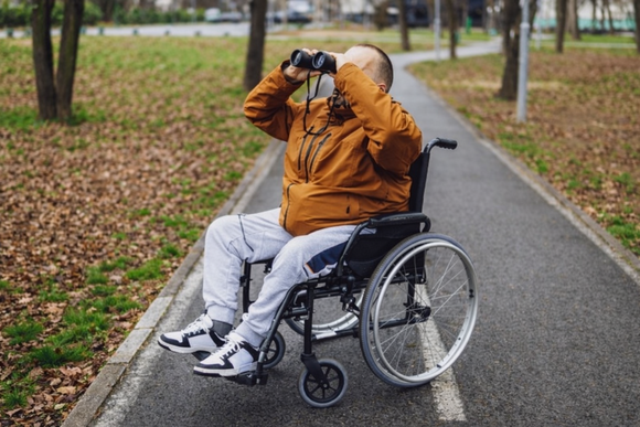 A man in a wheelchair on a paved trail looking through a pair of binoculars.