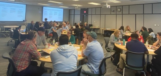 A diverse group of people sit at tables in a classroom. Screens on the walls show a presentation.