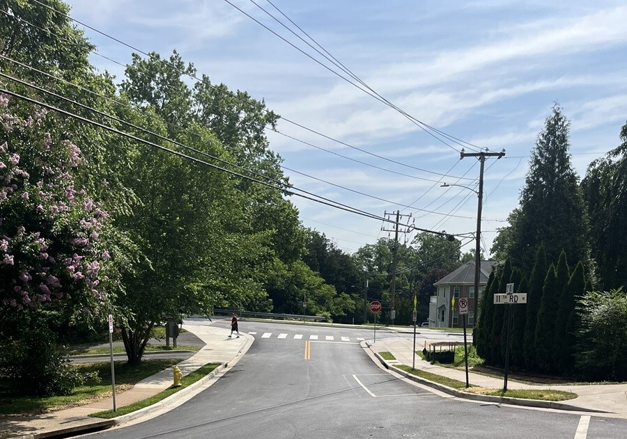 A street intersection with a marked crosswalk, sidewalks, overhead utility lines, trees, and a house in the background.