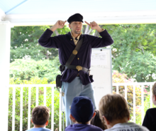 In instructor dressed in a civil war uniform is pointing to his hat while speaking to a group of children.