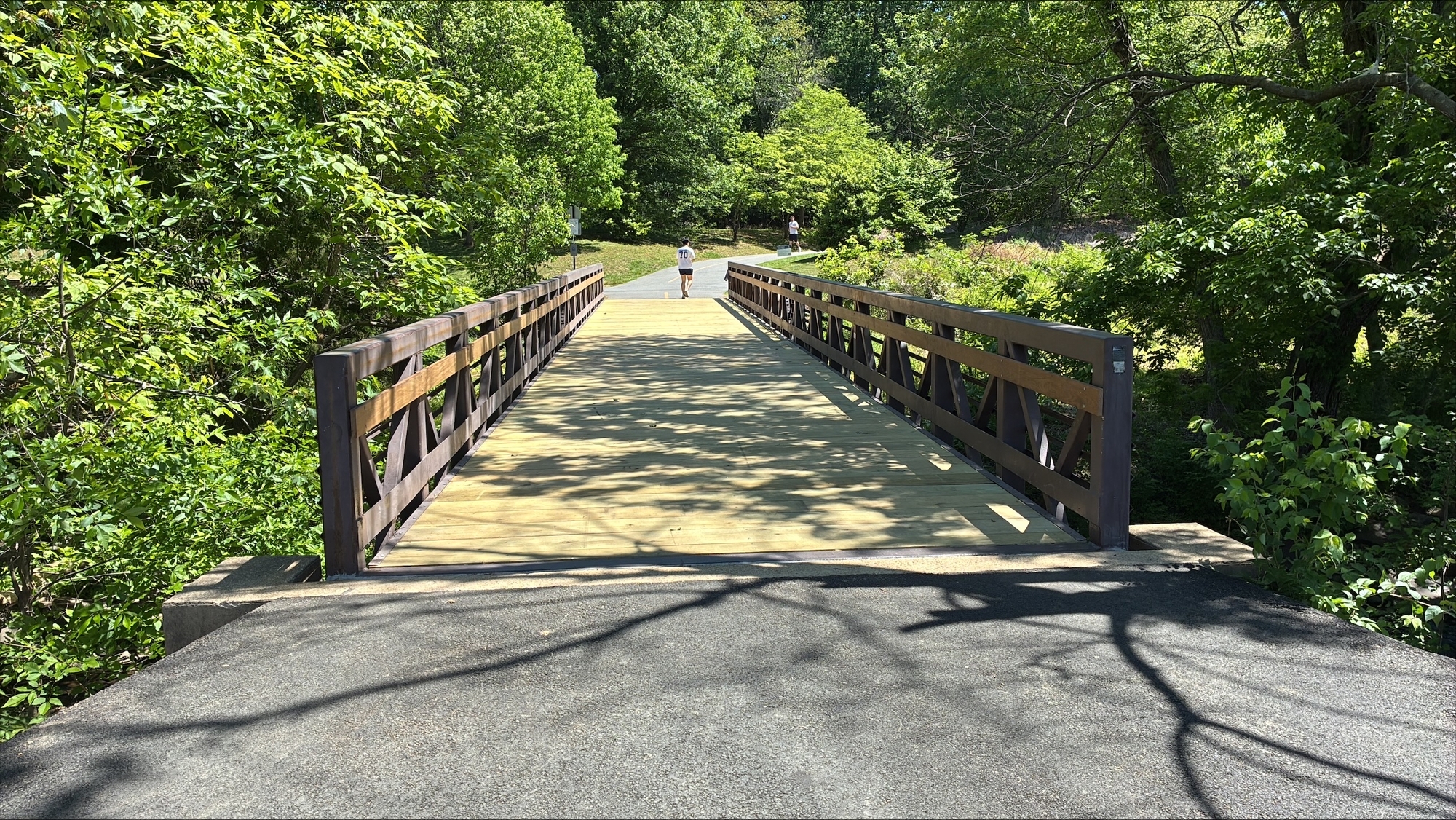 Sunlit wooden pedestrian bridge on a paved Bluemont trail, surrounded by dense green trees, with a person walking in the distance.