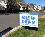 ID: A vision zero slow down sign on a neghborhood street, featuring the Cautionary Snail mascot forming the L and O in slow.
