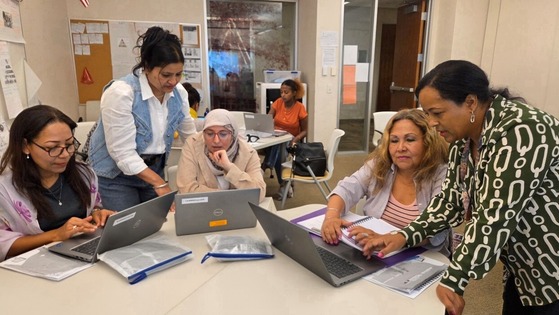A diverse group of Arlingtonians sits at a table with laptops. Two instructors stand nearby and assist the students.