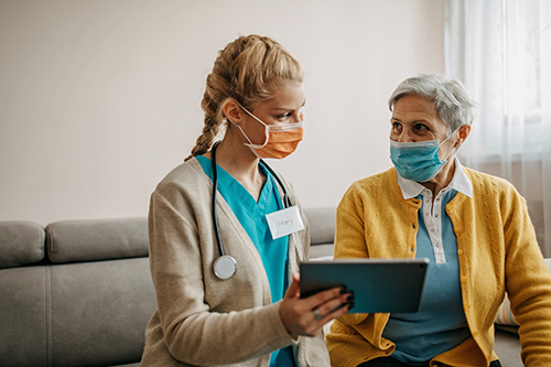 Photograph of medical provider sitting on a couch with a patient reviewing documentation on a tablet. Both are wearing mask. 