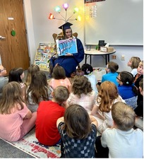 A senior student wearing cap and gown reading to young students