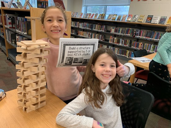 Two young female students pose with thumbs up next to a tower they built made of KEVA planks