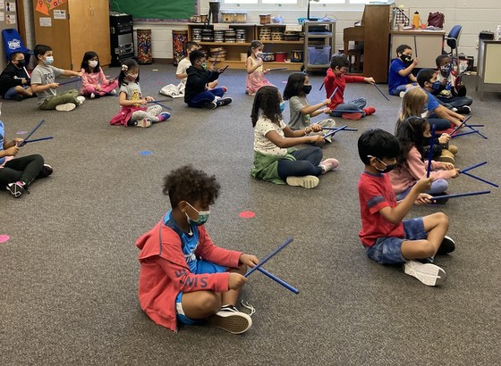 Photo of second grade students sitting on the floor playing rhythm sticks
