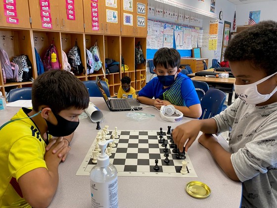 Photo of two students playing chess while being observed by a third.