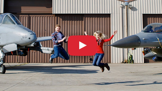 Two children jumping in the air next to fighter jets. 