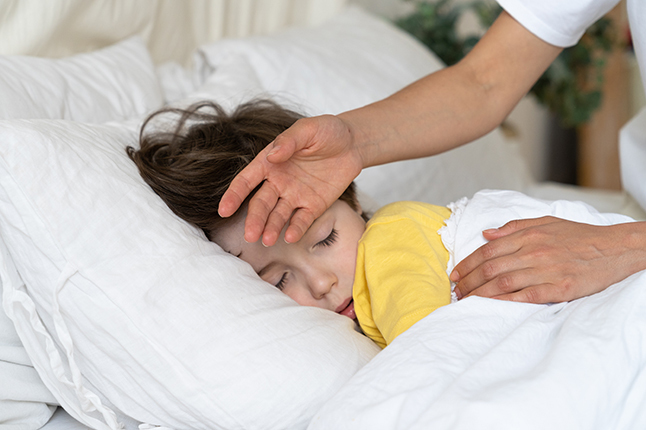 Young child in bed with a hand checking his forehead for a fever.