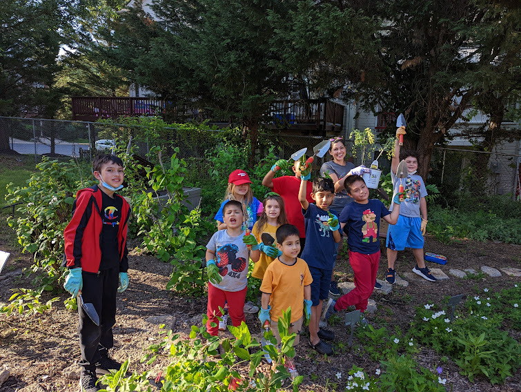 Photo of a group of students and adults outside near a garden. They are holding gardening tools and smiling.