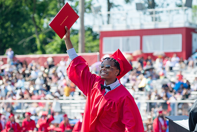 An FCPS Graduate raising their diploma and smiling.  Congratulations Fairfax County Public Schools 2022 Graduates