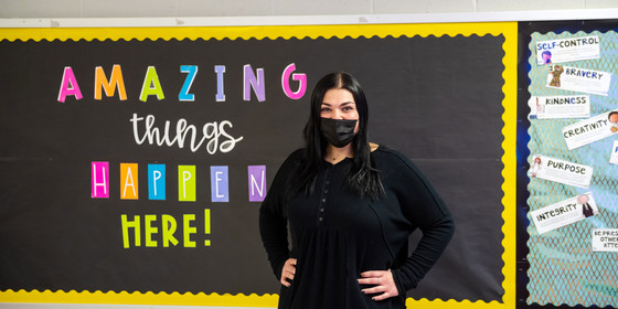 A teacher stands next to a bulletin board reading "Amazing things happen here!"