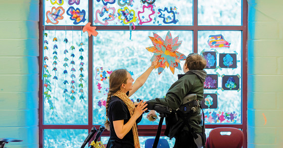 A teacher helps a student place a paper flower on a window.