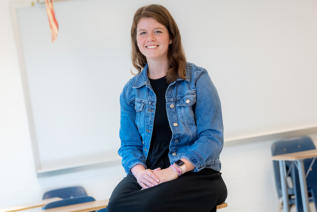A young teacher sits on a desk, smiling.