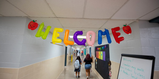 A balloon sign reads "welcome" over two students walking down the hall