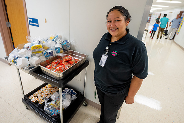 A cafeteria worker stands next to a cart of student breakfasts